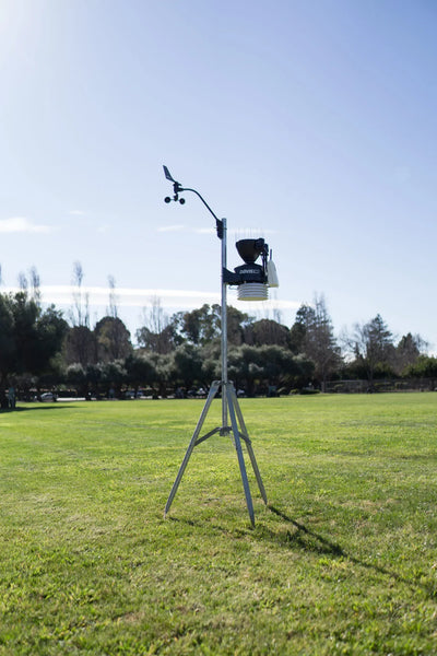 Weather station by Davis Instruments with anemometer, thermometer, rain collector on metal tripod in grassy field.
